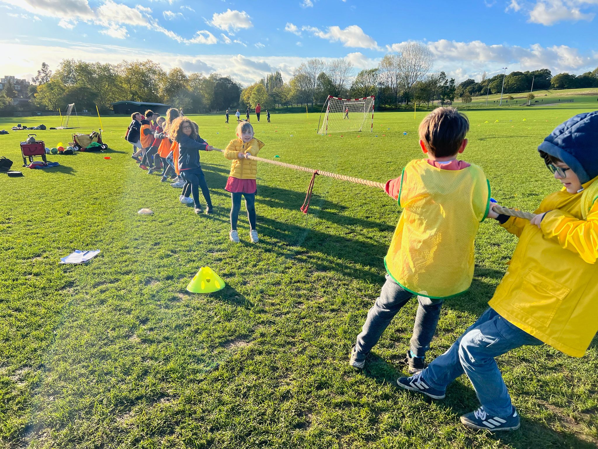 Photo of two groups of children wearing orange and yellow vests playing tug-of-war in a park on a sunny day