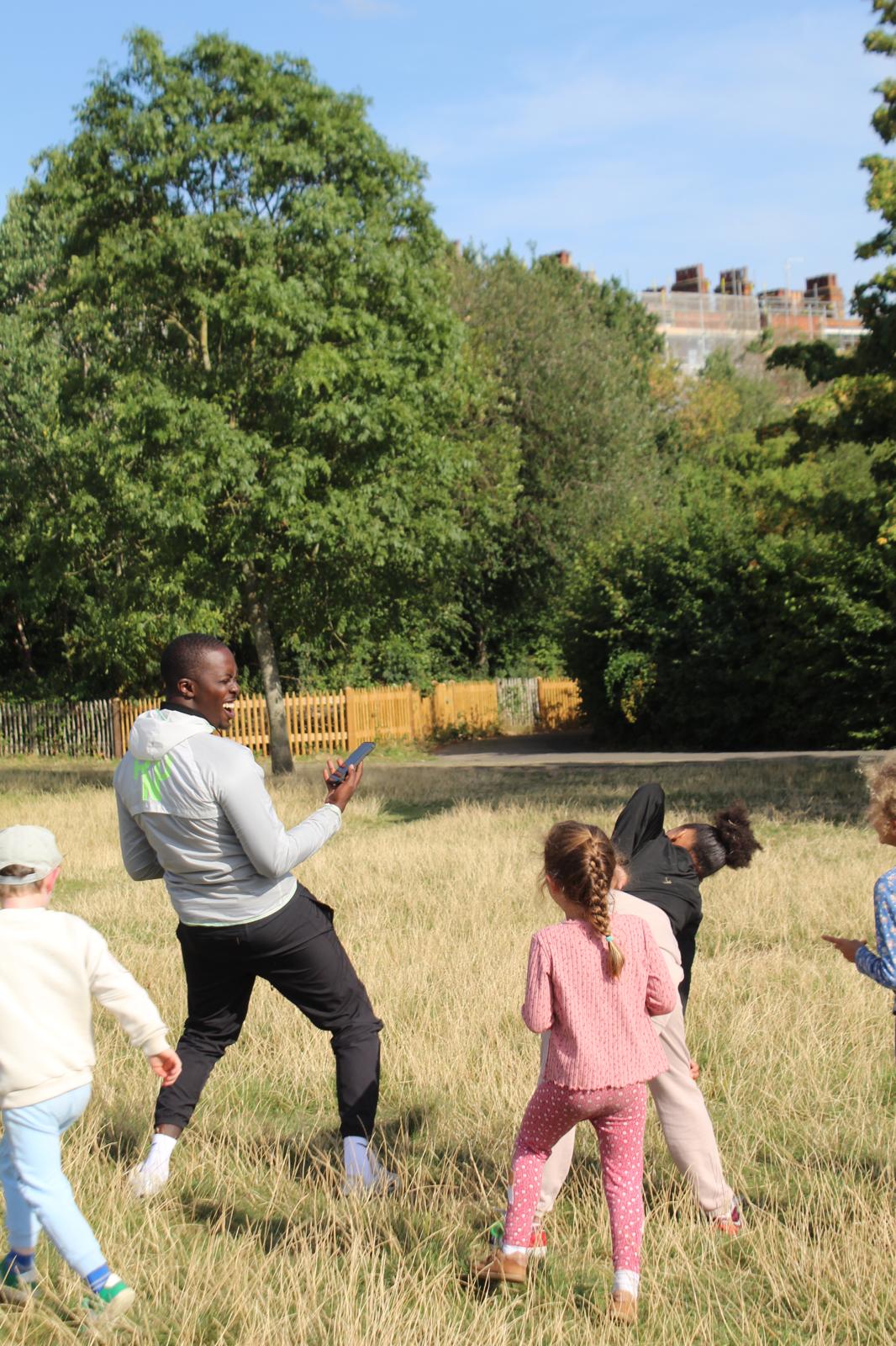 Photo of a coach laughing with children playing around him in a park on a sunny day